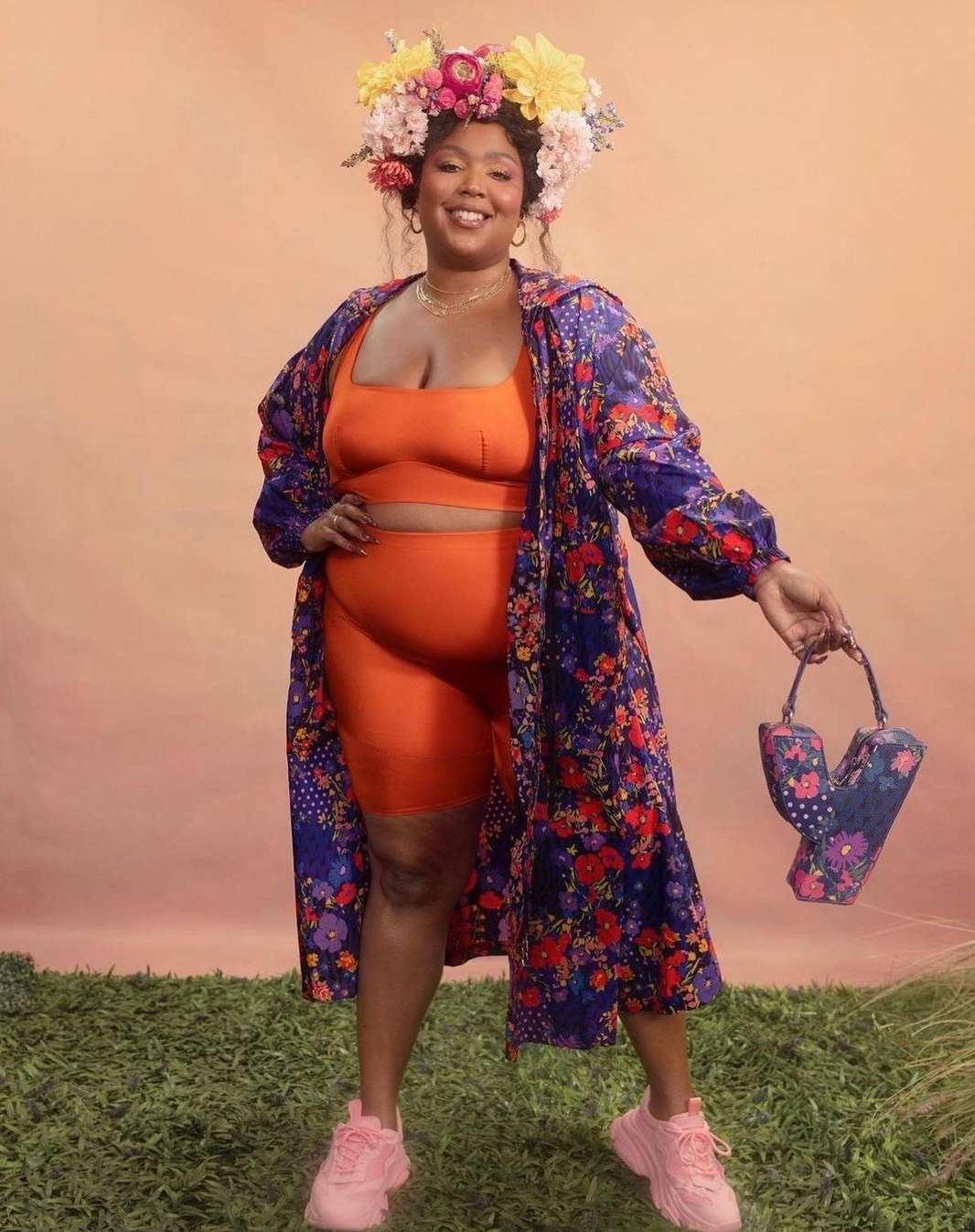 Woman with floral crown and floral-patterned robe poses outdoors against light peach backdrop with floral handbag and pink sneakers.