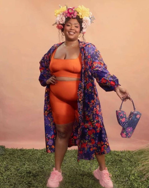 Woman with floral crown and floral-patterned robe poses outdoors against light peach backdrop with floral handbag and pink sneakers.