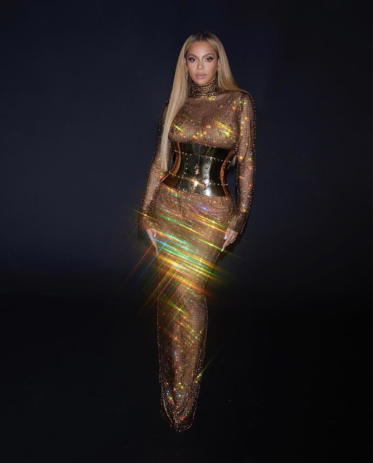 Beyoncé poses in a floor-length, golden chainmail dress with a metal corset on a dark background, radiating light and elegance.