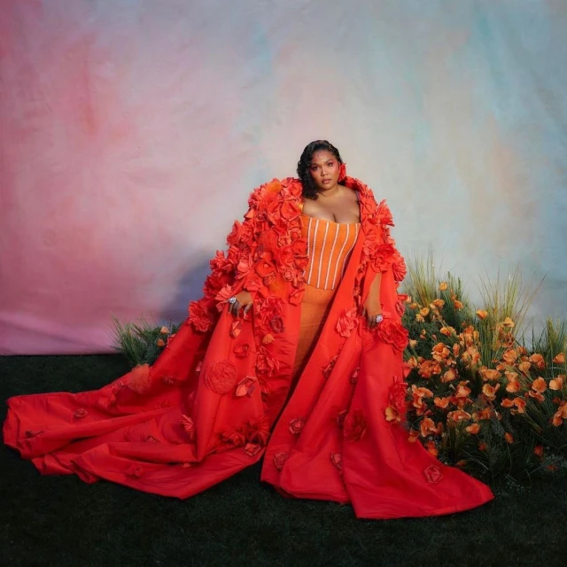 Lizzo poses in an orange dress and red floral coat with train in a photo studio with a colorful background and green grass.