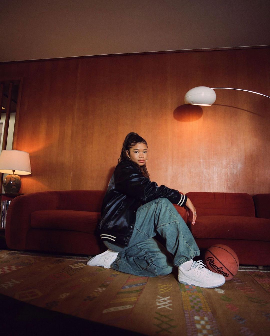 Woman in a black bomber jacket and baggy jeans kneeling in front of a red sofa in a room with wood-paneled walls.
