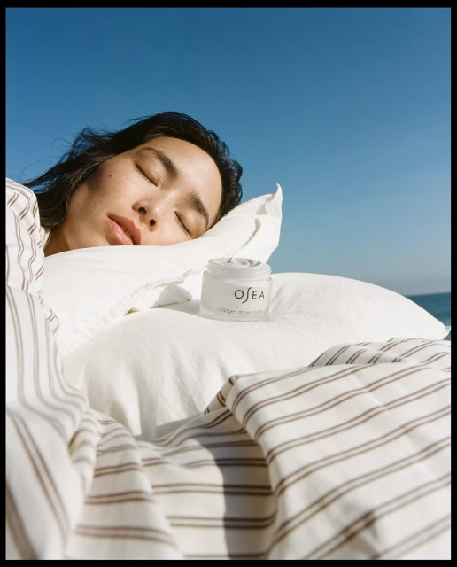 Asian woman sleeping outdoors with Osea collagen cream jar, striped linens, pillow, blue sky, and ocean background in a beauty product shot.