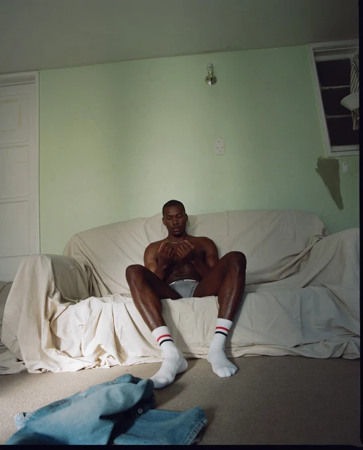 Shirtless man sits on a white couch in a room with light green walls, wearing socks and underwear. Jeans are on the floor.