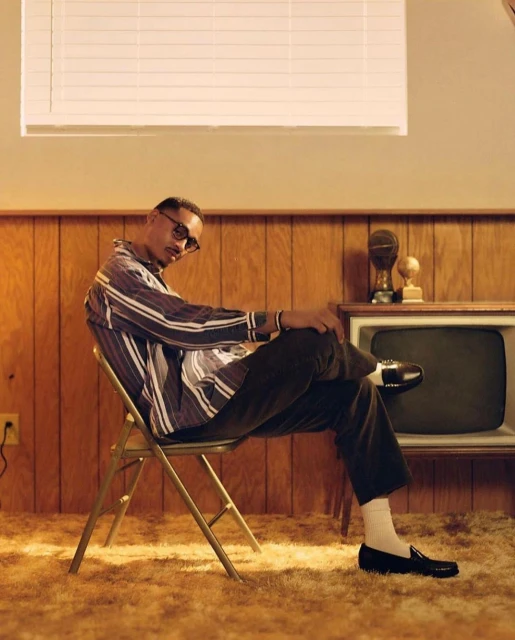 Man in striped shirt sits on chair, wearing glasses, with vintage TV and wood paneling in background, creating retro-style scene.
