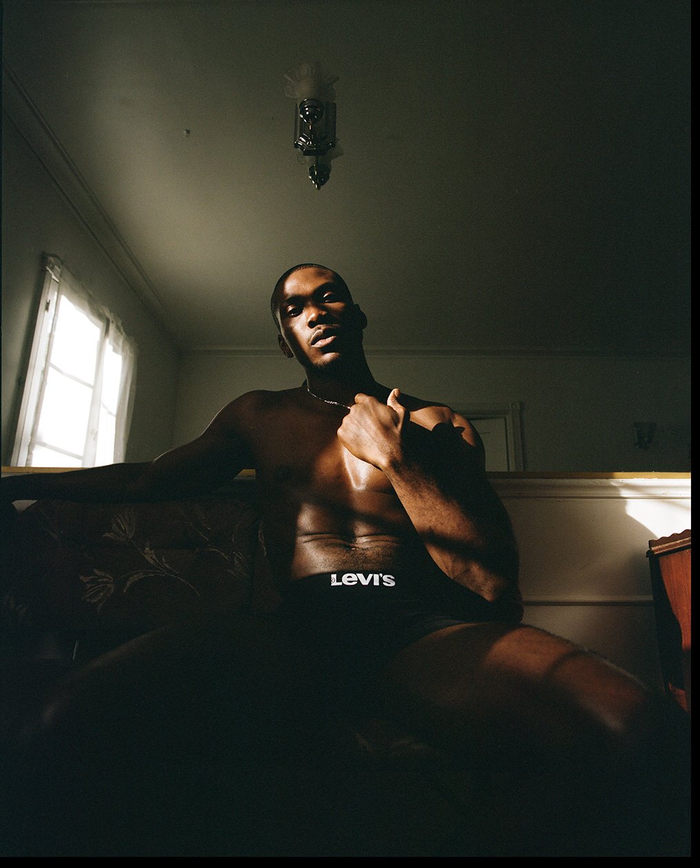 Shirtless black man seated in a dimly lit room wearing black underwear with white logo lettering, photographed by Robin Harper.