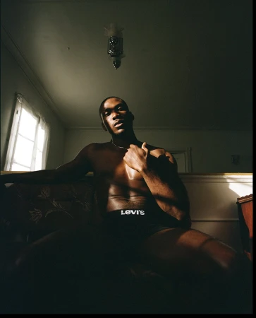 Shirtless black man seated in a dimly lit room wearing black underwear with white logo lettering, photographed by Robin Harper.