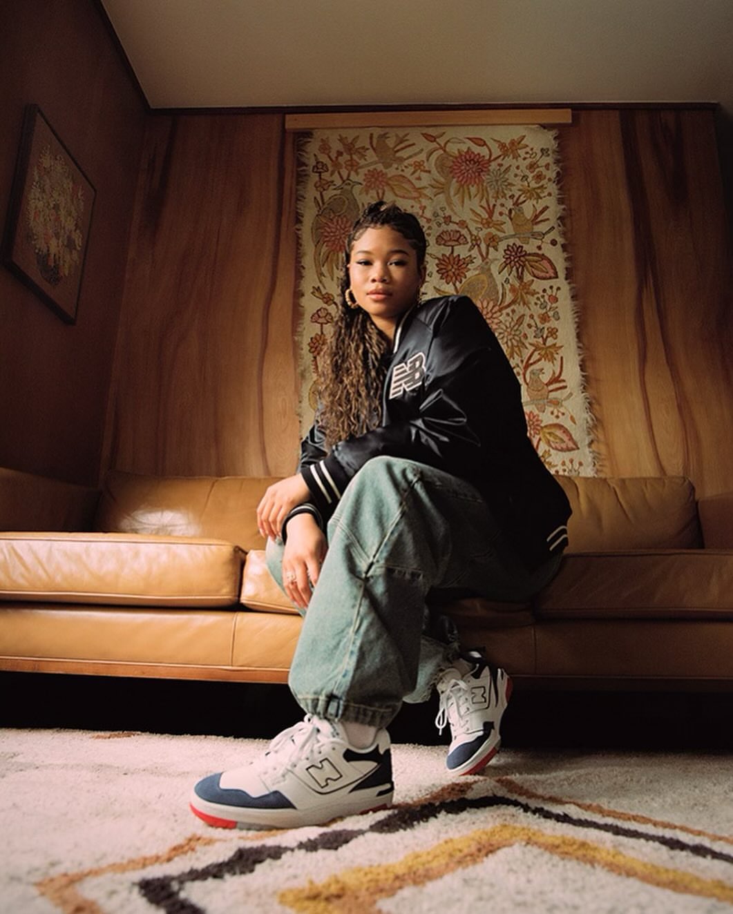 Athletic woman poses indoors wearing New Balance sneakers, blue jeans, and a varsity jacket. Soft indoor lighting and patterned carpet.