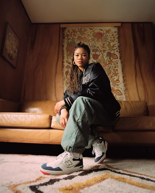 Athletic woman poses indoors wearing New Balance sneakers, blue jeans, and a varsity jacket. Soft indoor lighting and patterned carpet.