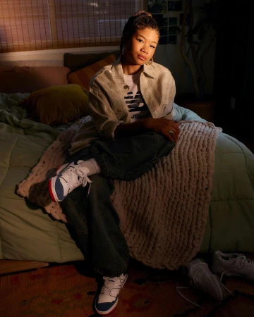 Portrait of a young woman seated on a bed in a softly lit bedroom, wearing a light patterned shirt and sneakers.