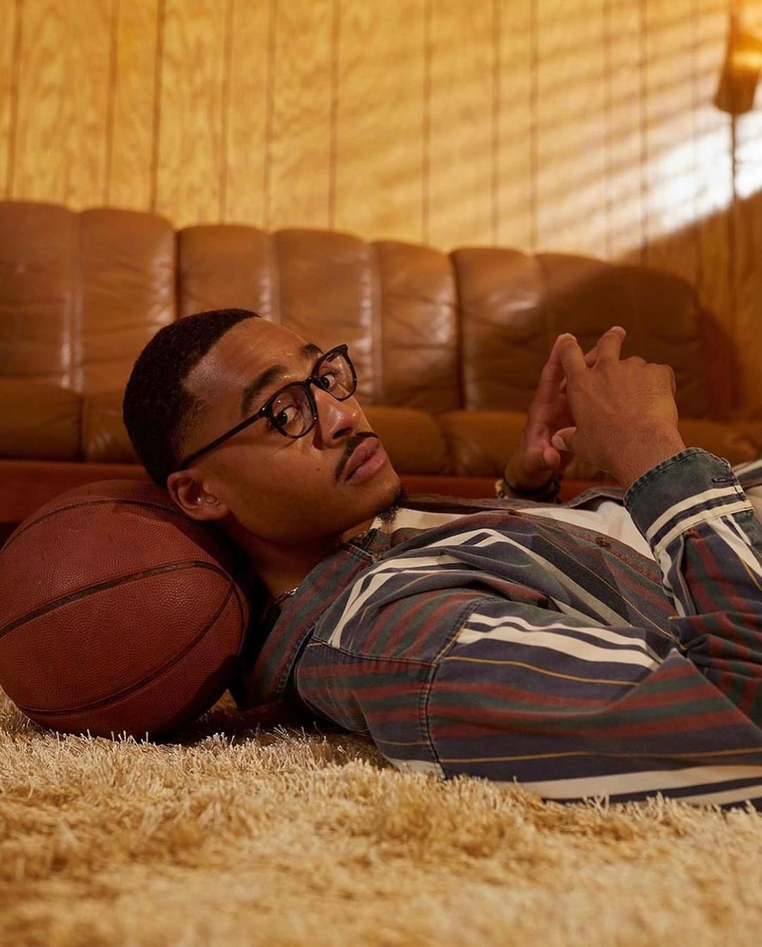 Young man with glasses lies on shag carpet with basketball, striped shirt, looking into the camera in an indoor room setting.
