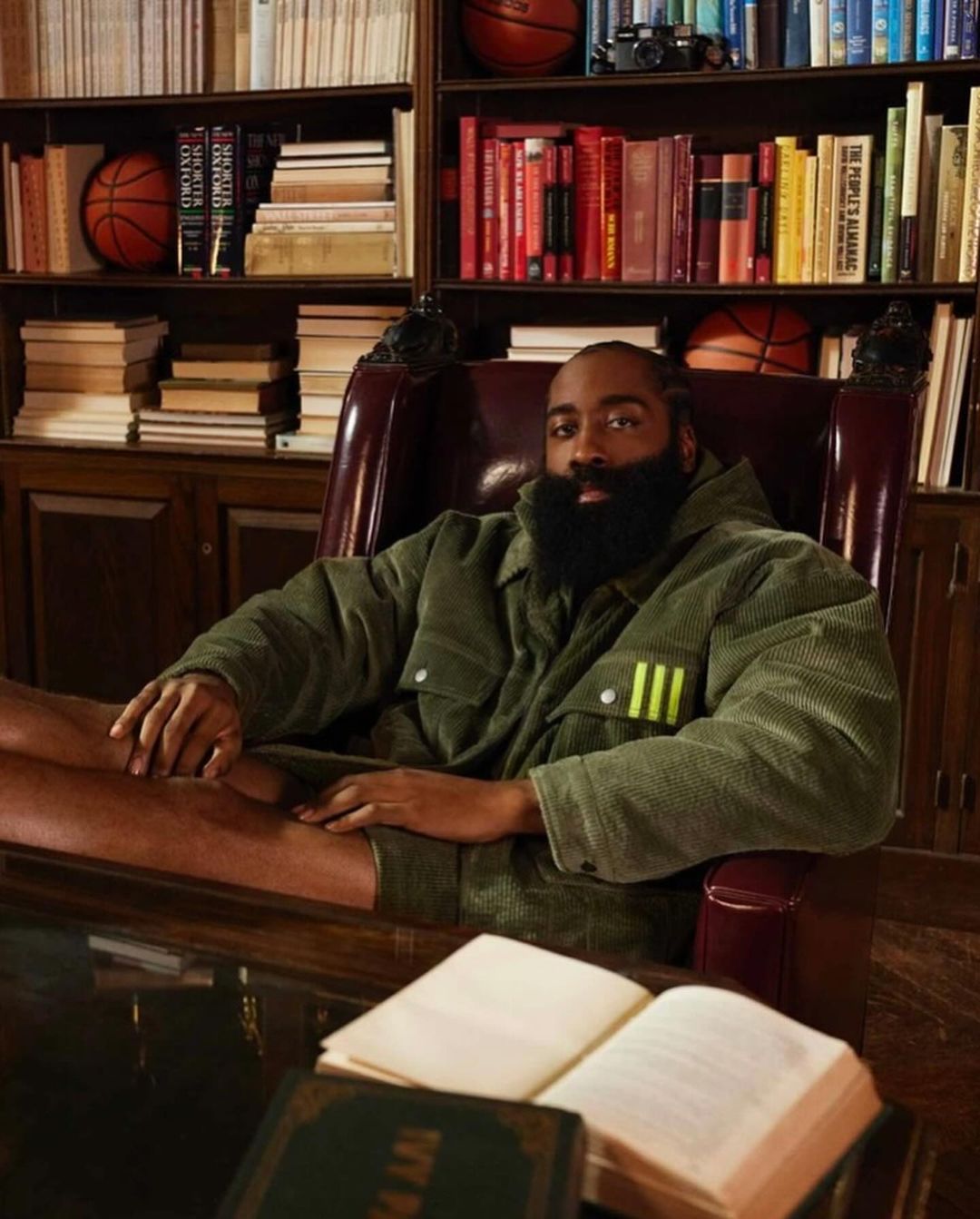 James Harden sits in a leather chair in an olive jacket, in front of a bookshelf with basketballs and books in a warm-toned room.