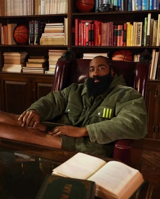 James Harden sits in a leather chair in an olive jacket, in front of a bookshelf with basketballs and books in a warm-toned room.