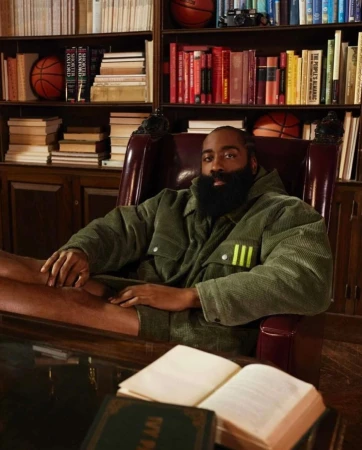 James Harden sits in a leather chair in an olive jacket, in front of a bookshelf with basketballs and books in a warm-toned room.