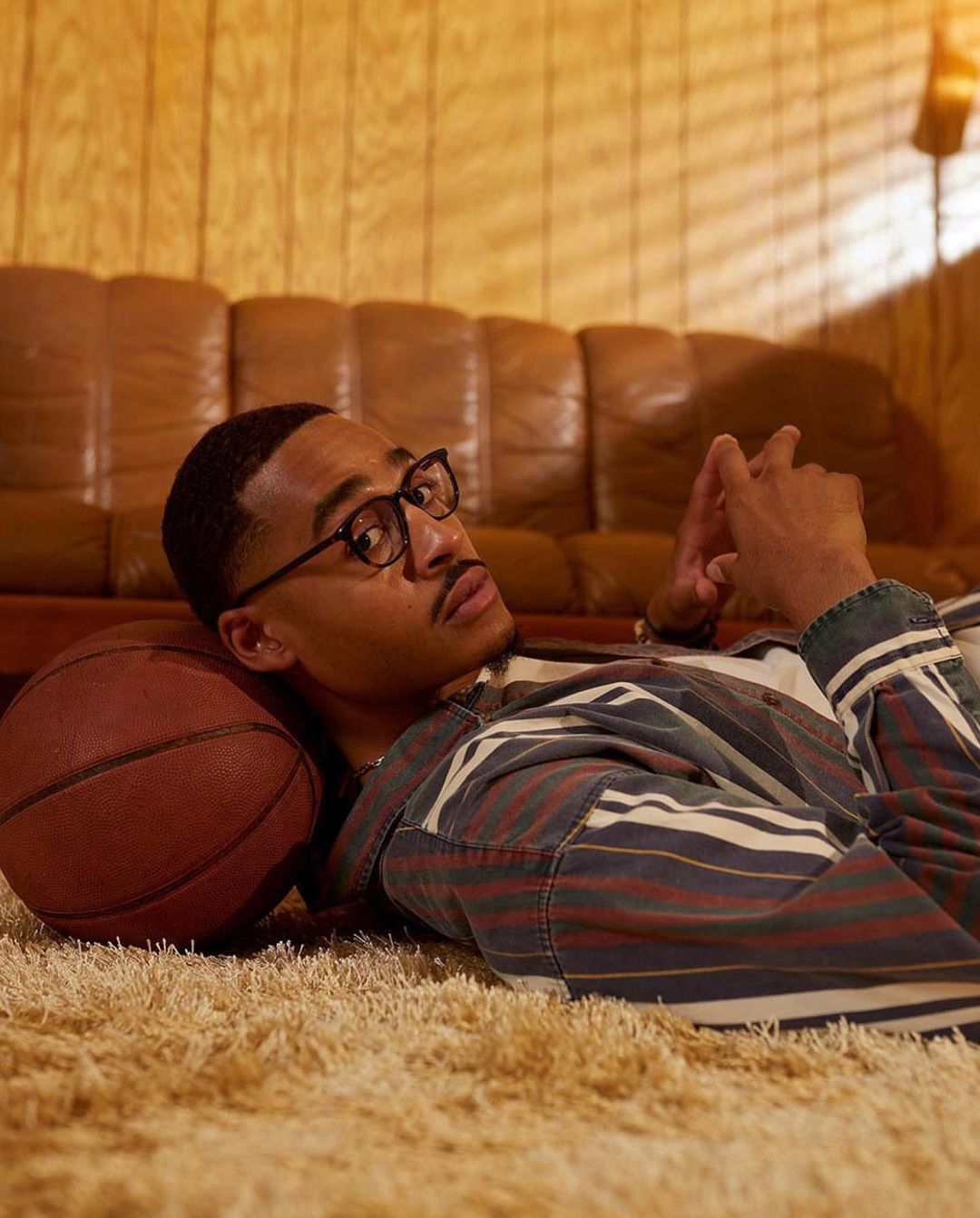 A young man with glasses lies on a shag carpet with a basketball, dressed in a striped jacket in a cozy, wood-paneled room.