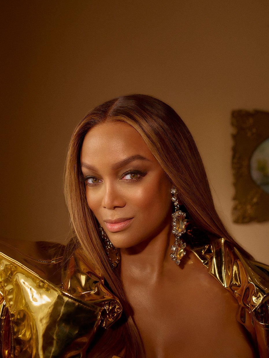 Glamorous headshot of Tyra Banks. She wears large chandelier earrings and a shiny gold jacket, posing against a neutral brown background.