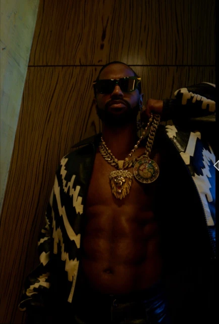 Man with exposed torso wearing a jacket, sunglasses, and large gold necklaces against a textured, dark background. Bold fashion statement.
