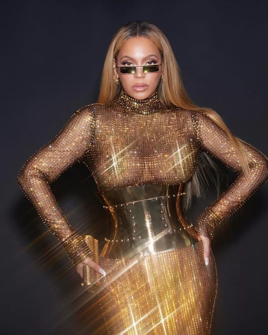 Beyonce poses in a glittering gold gown and rectangular sunglasses against a dark backdrop, showcasing a blend of fashion and celebrity.