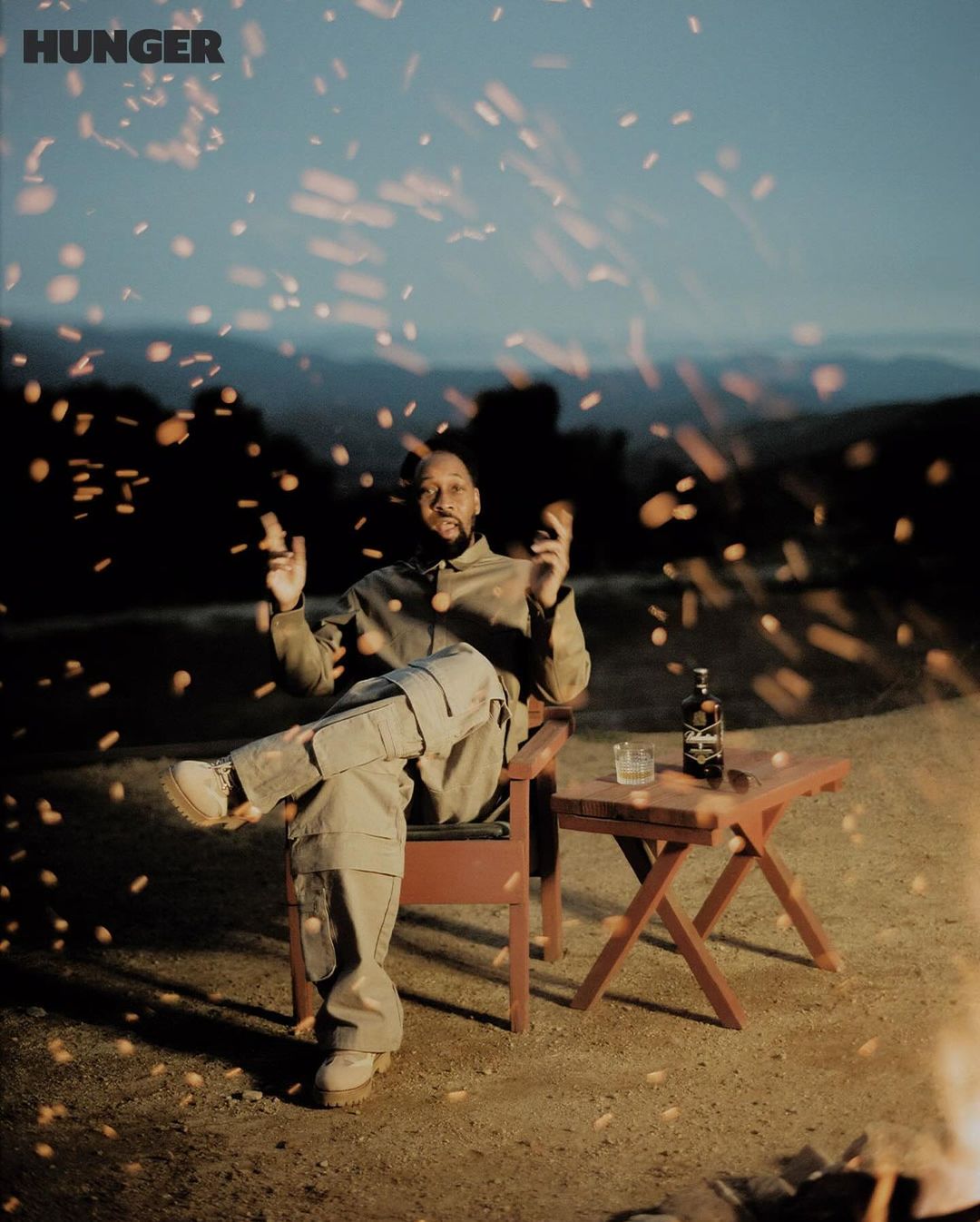 Portrait of a man sitting on a chair outside at night with a bottle and glass on a table, with visible sparks or bokeh.