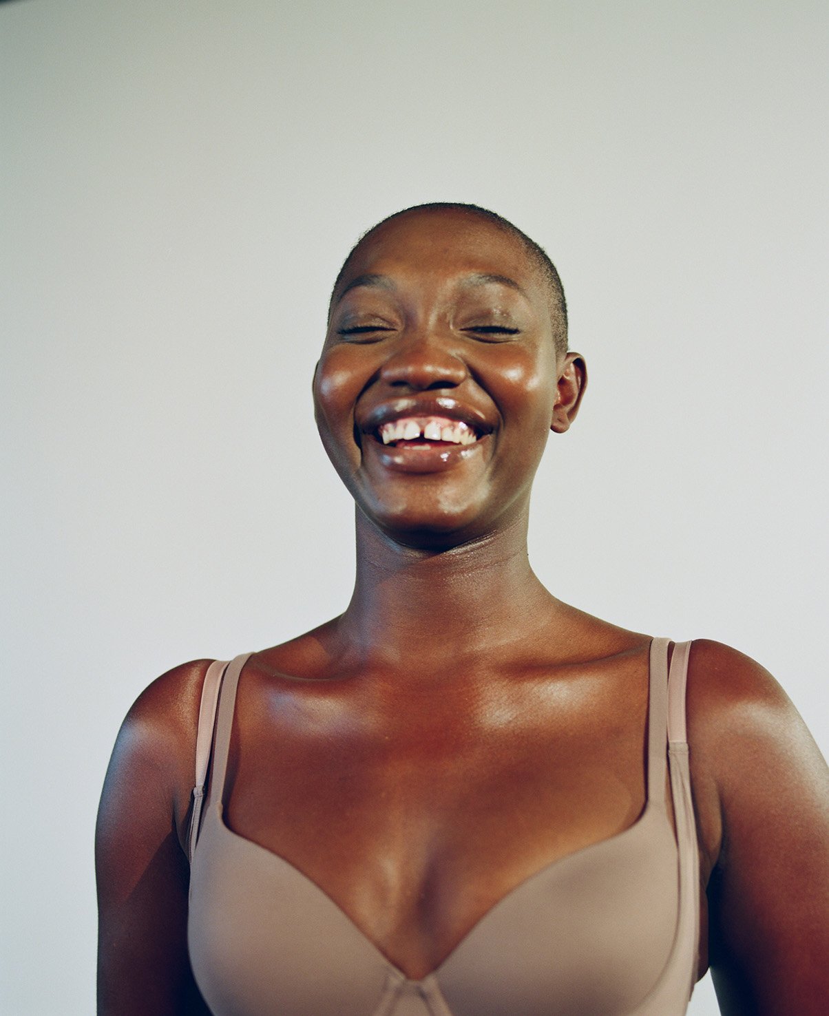 Close-up portrait of a smiling African-American woman with a shaved head, wearing a bra against a plain backdrop in a photo studio.