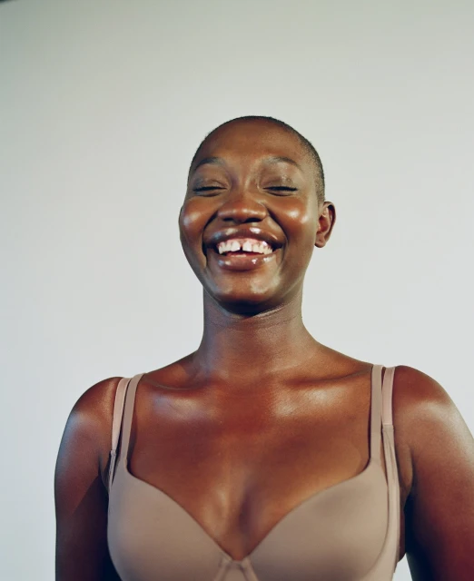 Close-up portrait of a smiling African-American woman with a shaved head, wearing a bra against a plain backdrop in a photo studio.