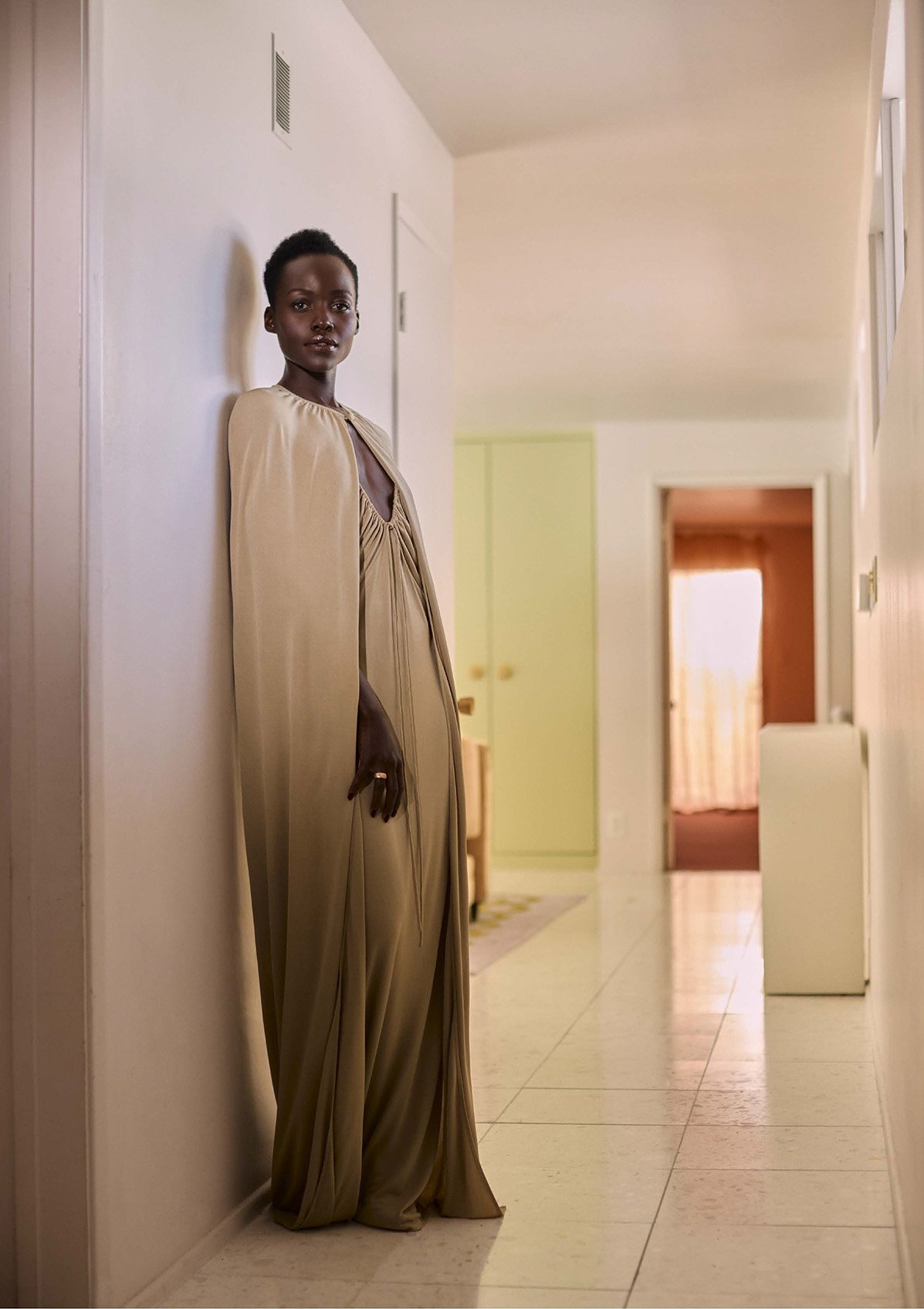 Black woman wearing a long gold hooded gown leaning against a white hallway wall in a Los Angeles interior by photographer Robin Harper