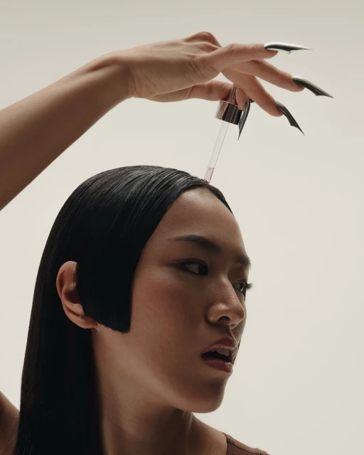 Elegant woman with sleek hair and long nails receives beauty treatment from dropper in a softly lit, neutral studio setting.