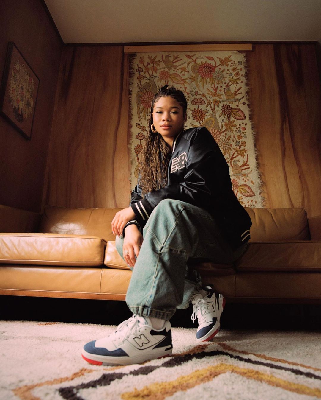 Young woman with curly hair poses on a sofa in a black jacket and jeans with white New Balance sneakers in an indoor setting.