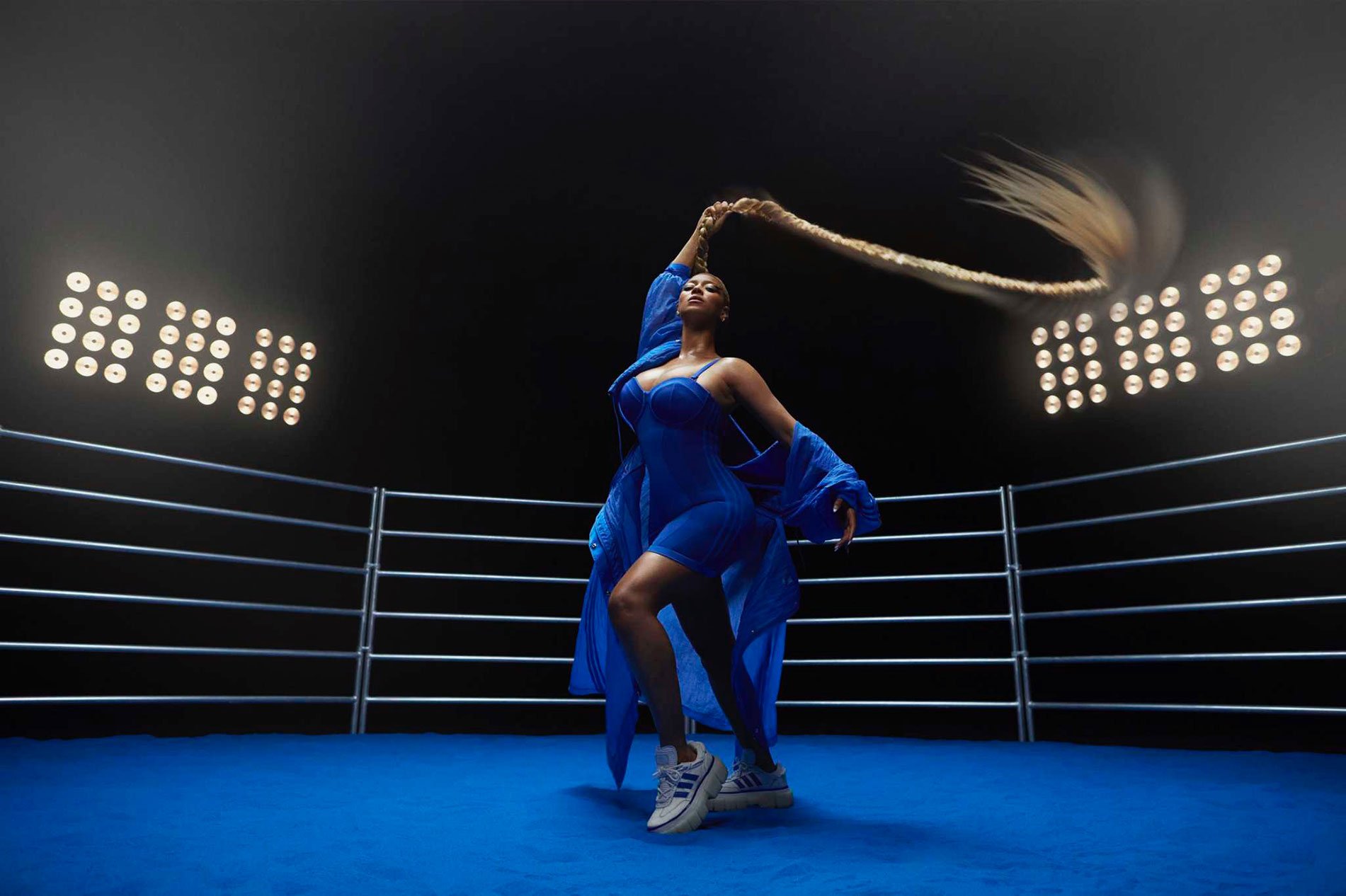 Woman in blue outfit poses in boxing ring under bright lights, holding long braid above her head, creating a dynamic and visually striking image.