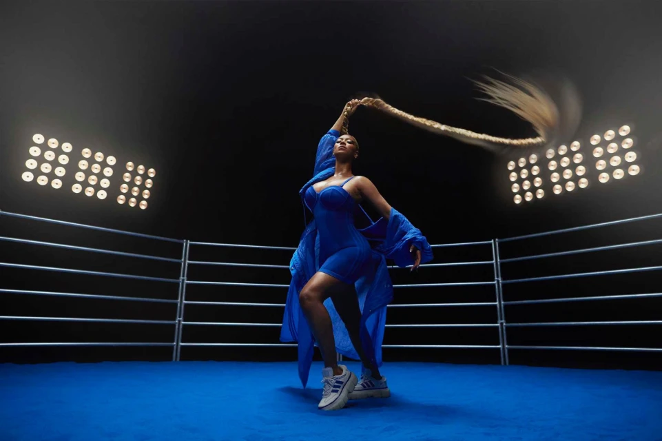 Woman in blue outfit poses in boxing ring under bright lights, holding long braid above her head, creating a dynamic and visually striking image.