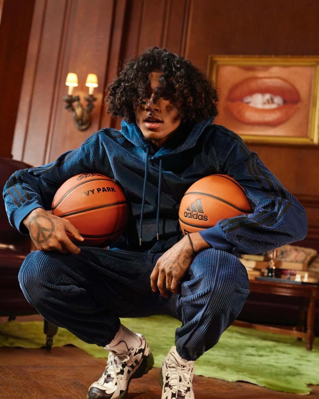 A young, dark-haired male model in a blue Adidas sweatsuit is squatting while holding two orange basketballs in an indoor setting.