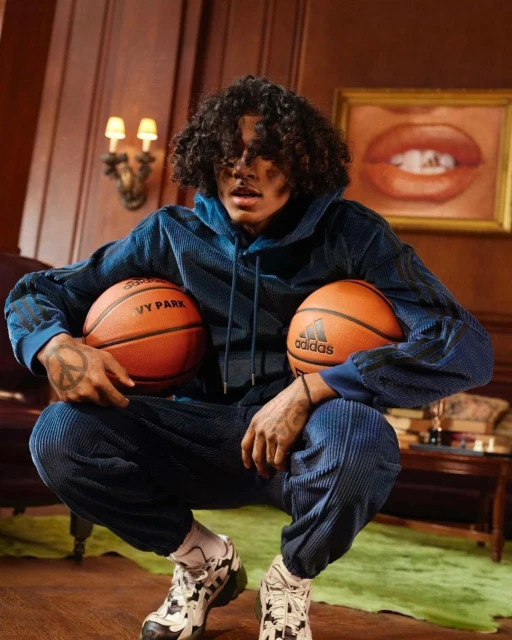 A young, dark-haired male model in a blue Adidas sweatsuit is squatting while holding two orange basketballs in an indoor setting.