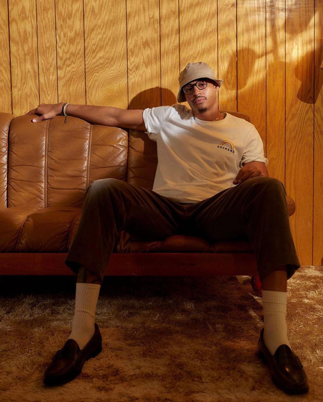 Man in white t-shirt and bucket hat seated on a brown leather couch against wood paneling