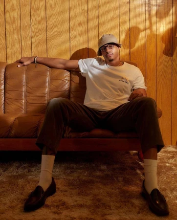 Man in white t-shirt and bucket hat seated on a brown leather couch against wood paneling