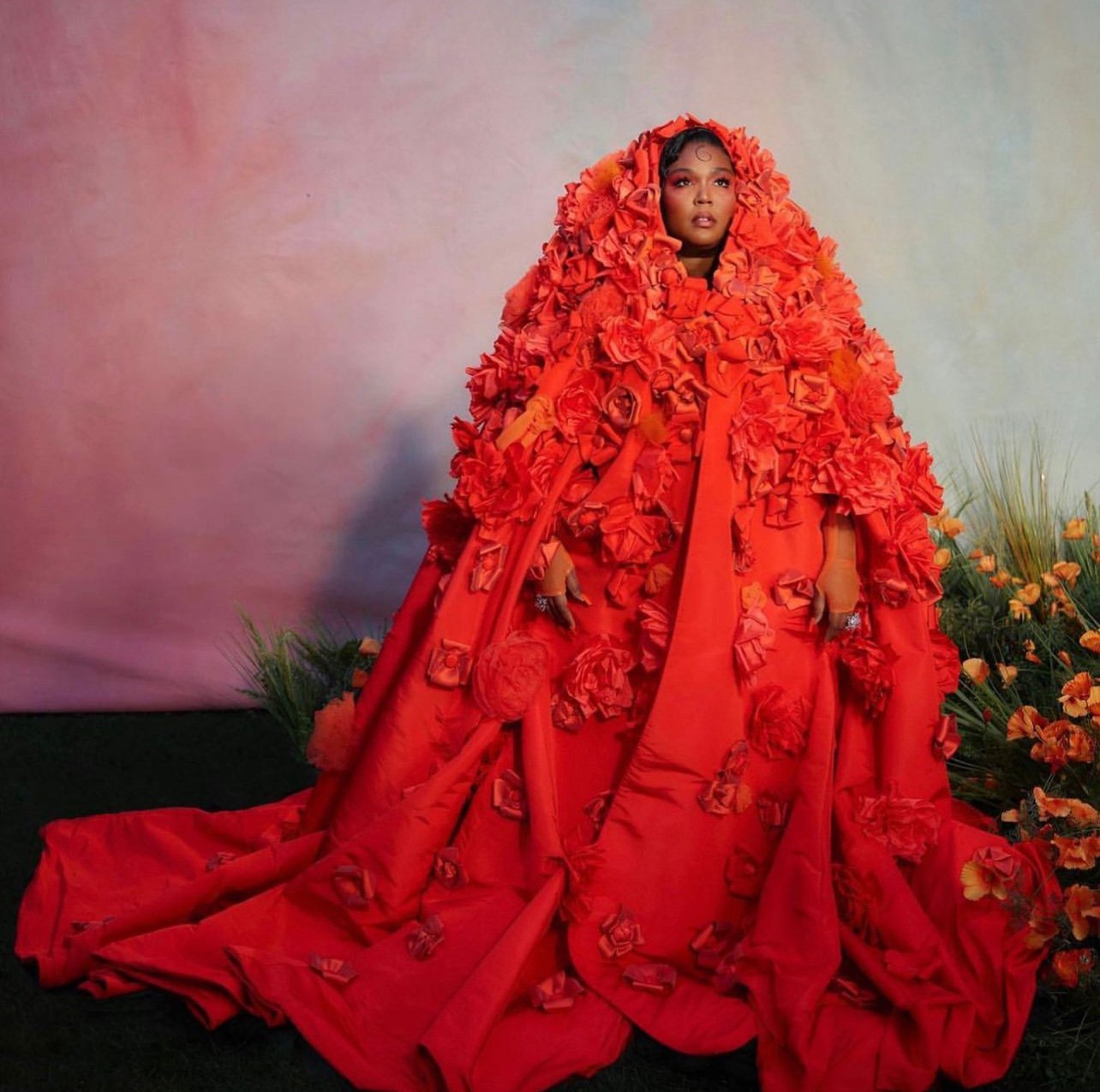 A person posing in a vibrant red gown adorned with floral details, against a colorful backdrop with floral accents.