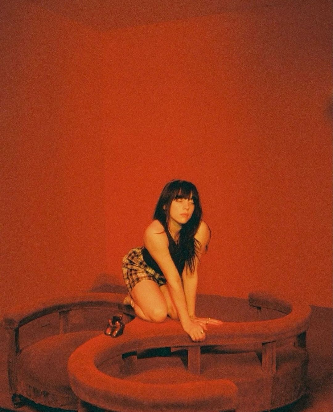 A woman kneels on red furniture in a photo studio with a solid red backdrop, wearing a black top and plaid shorts in a close-up.