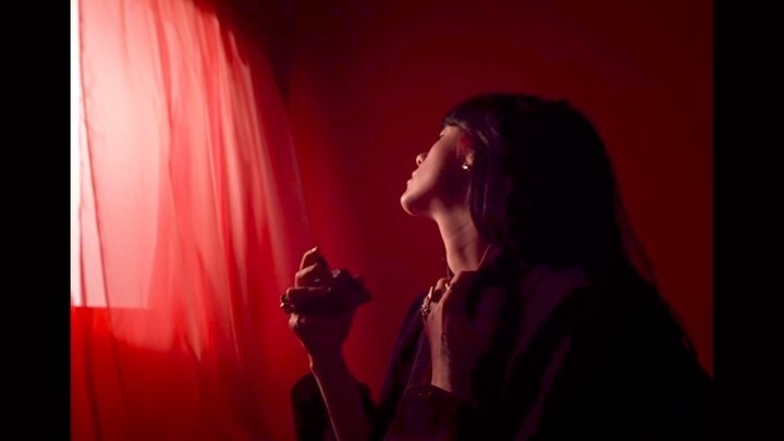 Young woman sprays perfume in a dimly lit, atmospheric setting with red curtains creating a dramatic, sensual ambiance.