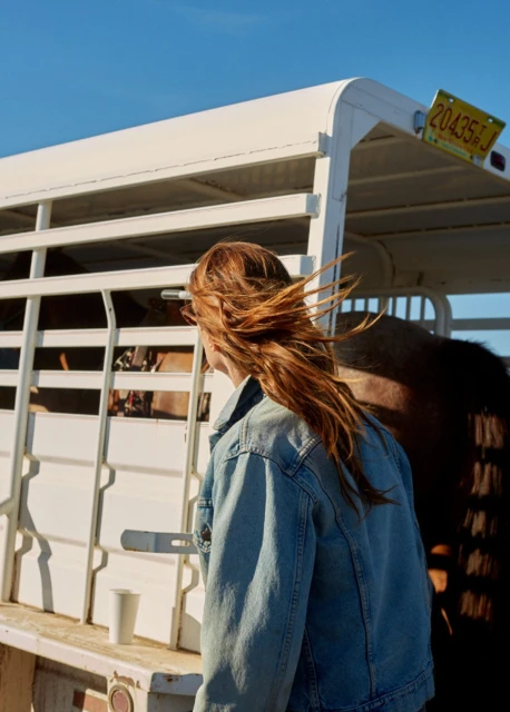 Adult female with long red hair standing next to a white horse trailer on a sunny day with a blue sky in a rural environment.