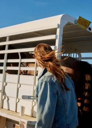 Adult female with long red hair standing next to a white horse trailer on a sunny day with a blue sky in a rural environment.