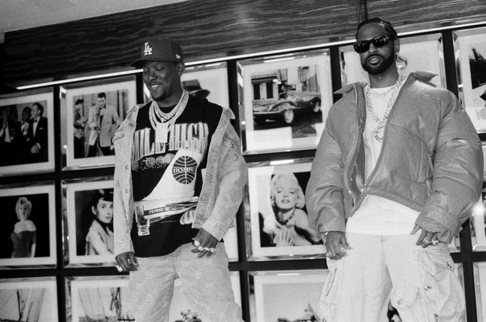 Two adult men pose in streetwear style in a photo studio with framed pictures on the wall. One man wears a baseball cap and denim jacket.
