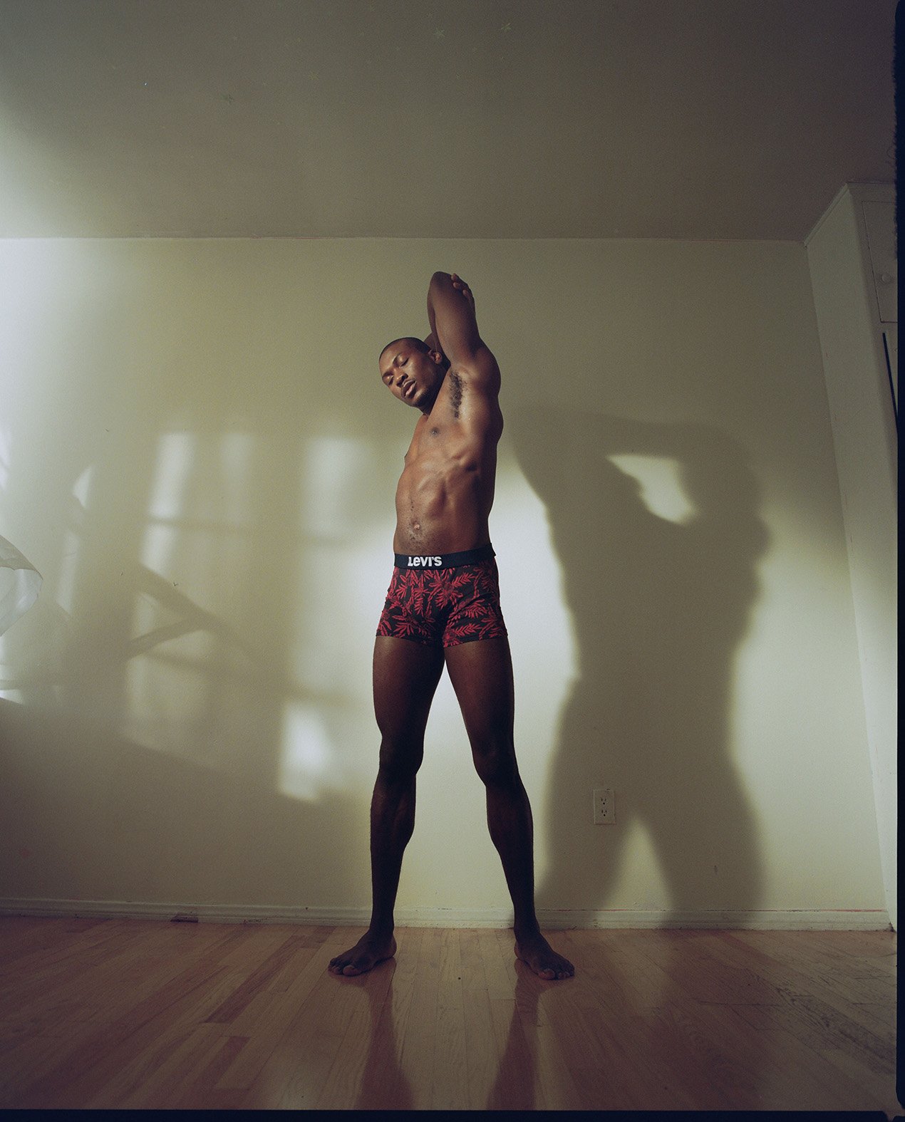 Black man stretching in patterned underwear with arms raised inside a room with light wooden floors.