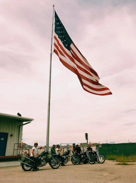 Group of adult men on motorcycles beneath a waving American flag in an outdoor, daytime setting with a building nearby.