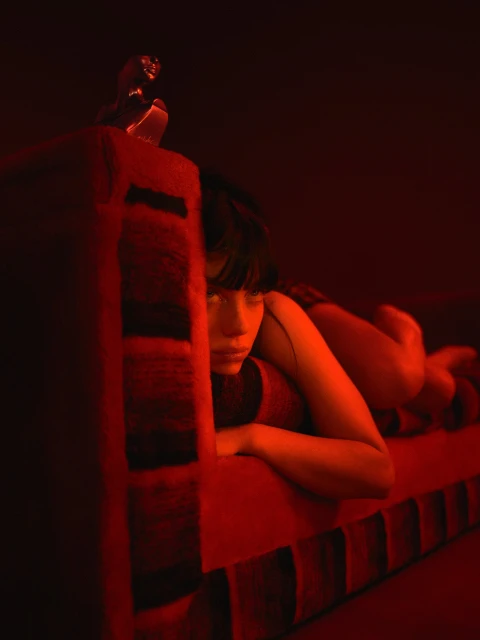 An adult female with bangs and dark hair lounges on a red-toned upholstered sofa in a dimly lit interior space, looking off-camera.