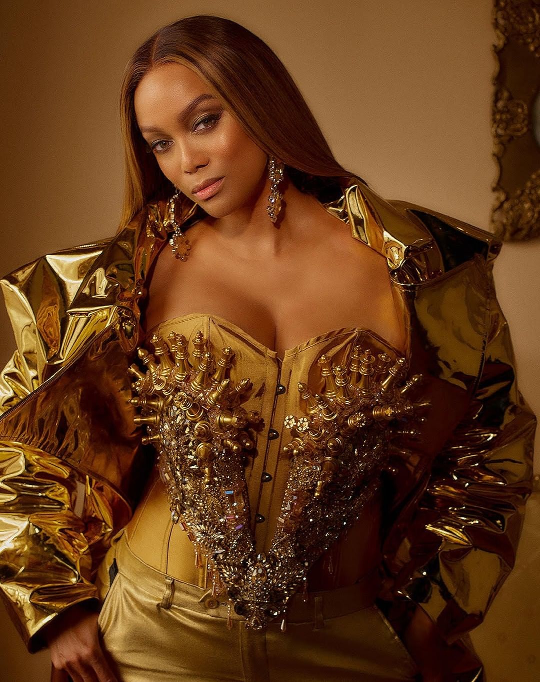 Tyra Banks poses in a gold corset top and matching jacket against a neutral backdrop with her hair parted on the side.