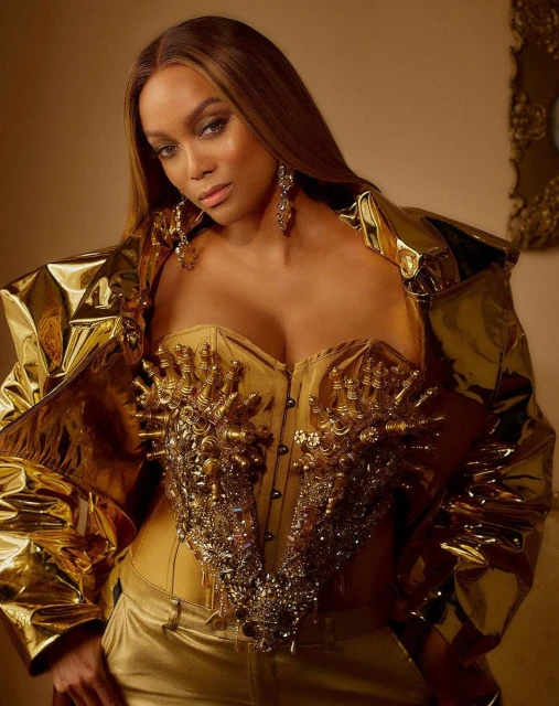 Tyra Banks poses in a gold corset top and matching jacket against a neutral backdrop with her hair parted on the side.