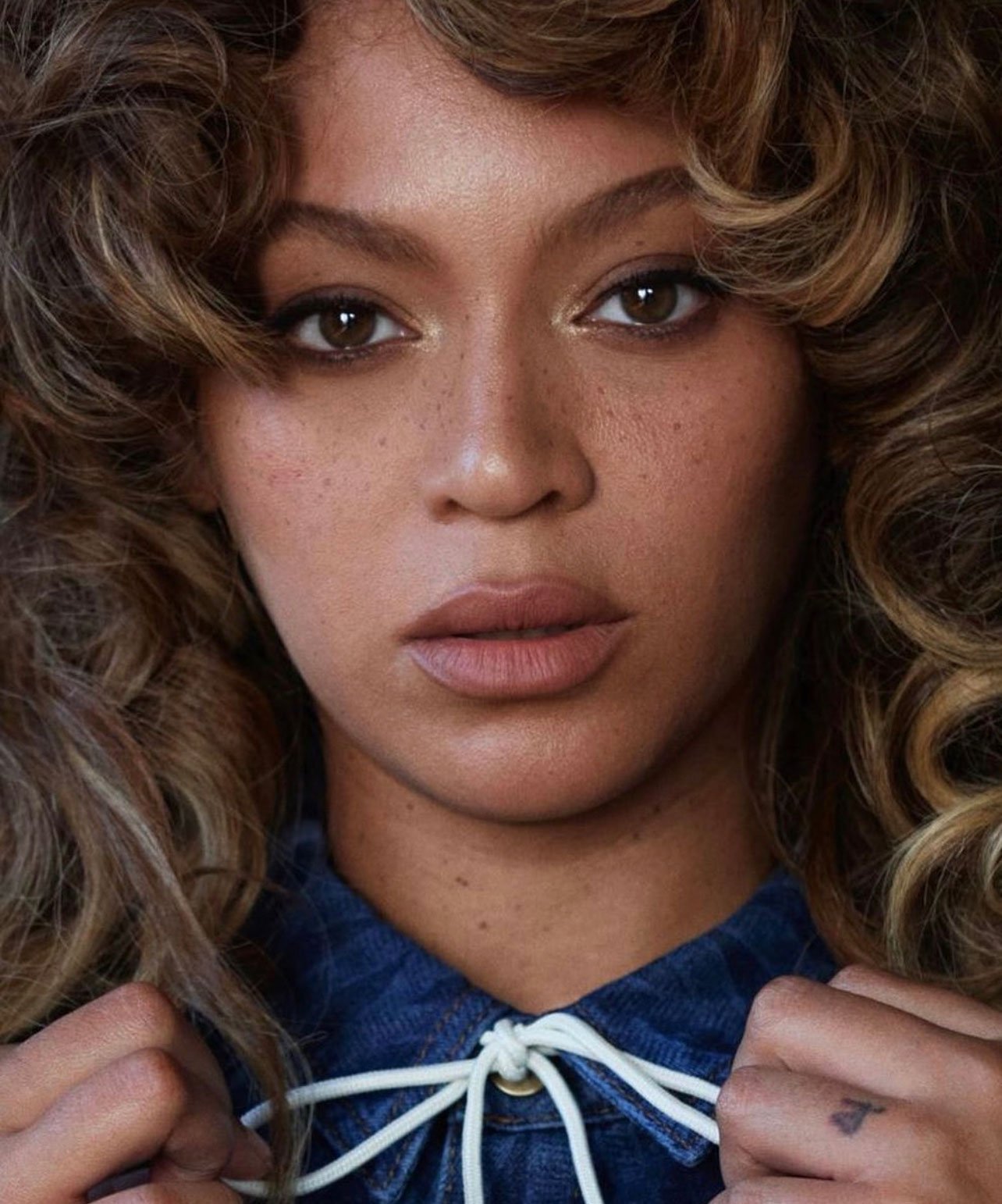 Close-up portrait of Beyoncé with curly blonde-highlighted hair wearing a blue denim top with white drawstrings, photographed by Robin Harper.