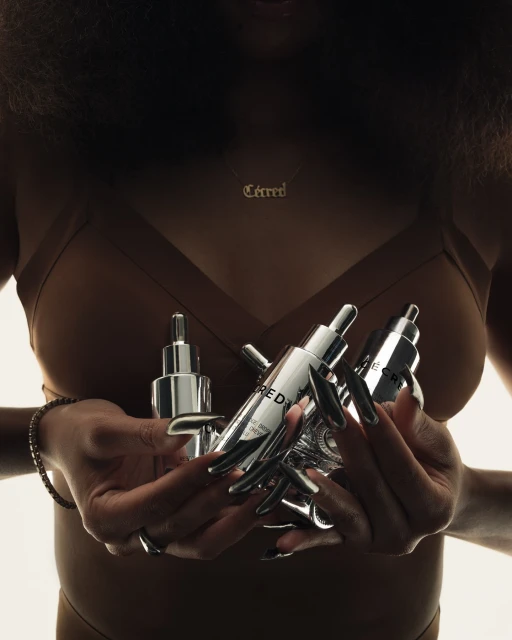 Woman holds mirrored beauty product bottles in hands with long nails. Dark hair, gold necklace, and brown outfit in a bright studio.
