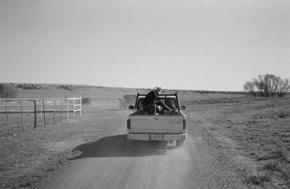 A black and white photo captures a vintage pickup truck driving on a dirt road through a rural landscape, carrying people in the truck bed.