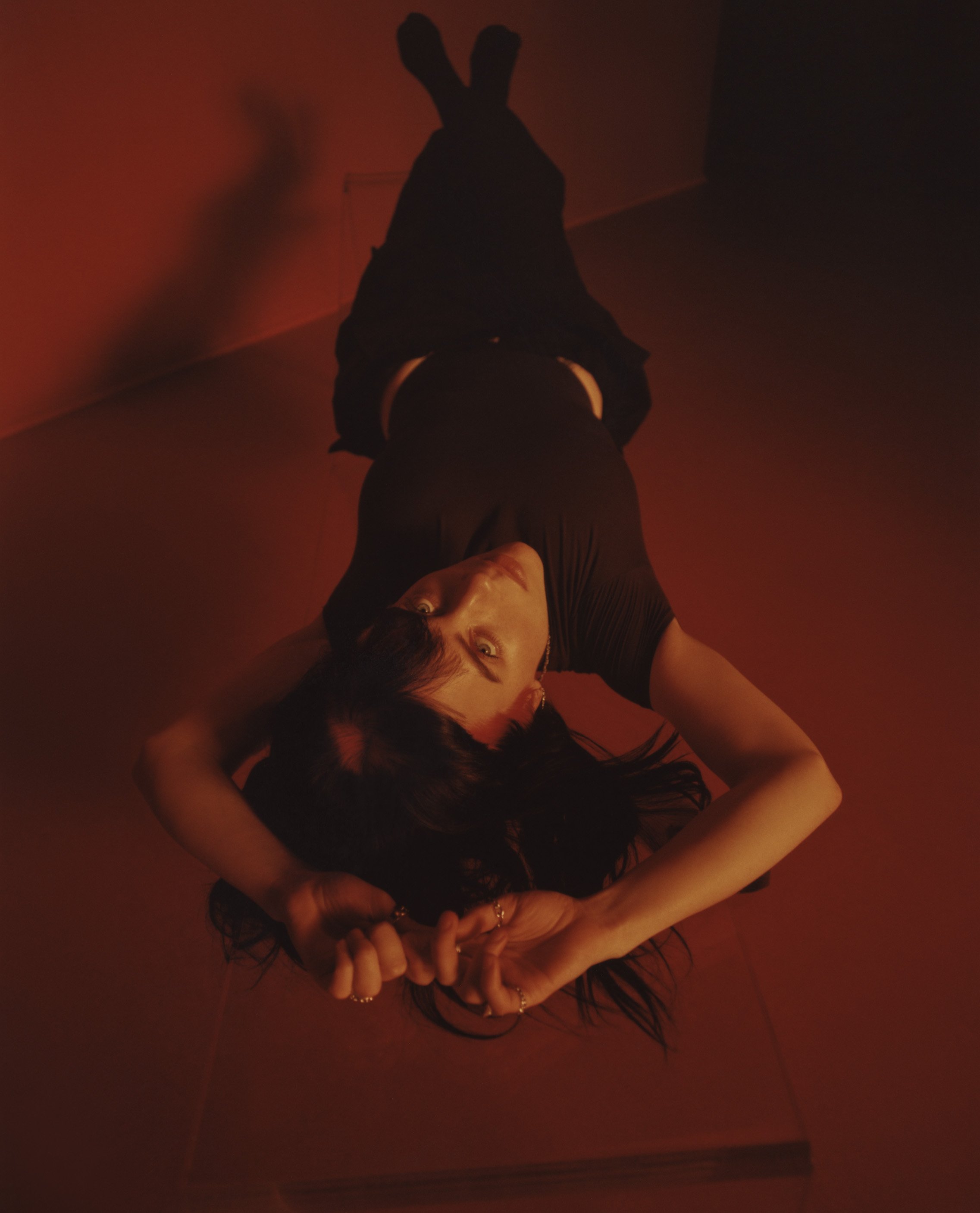 Woman in black lies on floor, shot from above. Dark hair surrounds her head as she looks up at the camera. Red walls and floor.