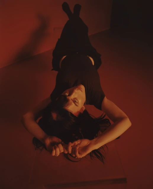 Woman in black lies on floor, shot from above. Dark hair surrounds her head as she looks up at the camera. Red walls and floor.