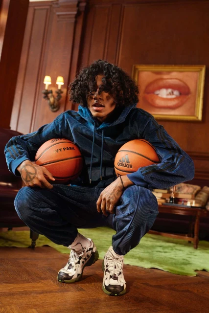 A person with curly hair wearing a blue corduroy hooded jumpsuit, holding two basketballs, and squatting on a wood floor in a room.